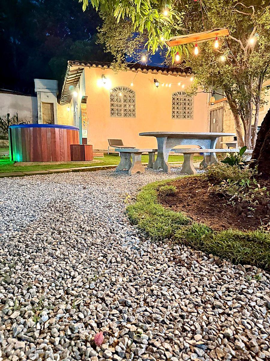 Área gourmet iluminada à noite, com mesa de pedra, spa de hidromassagem em ofurô de madeira e jardim